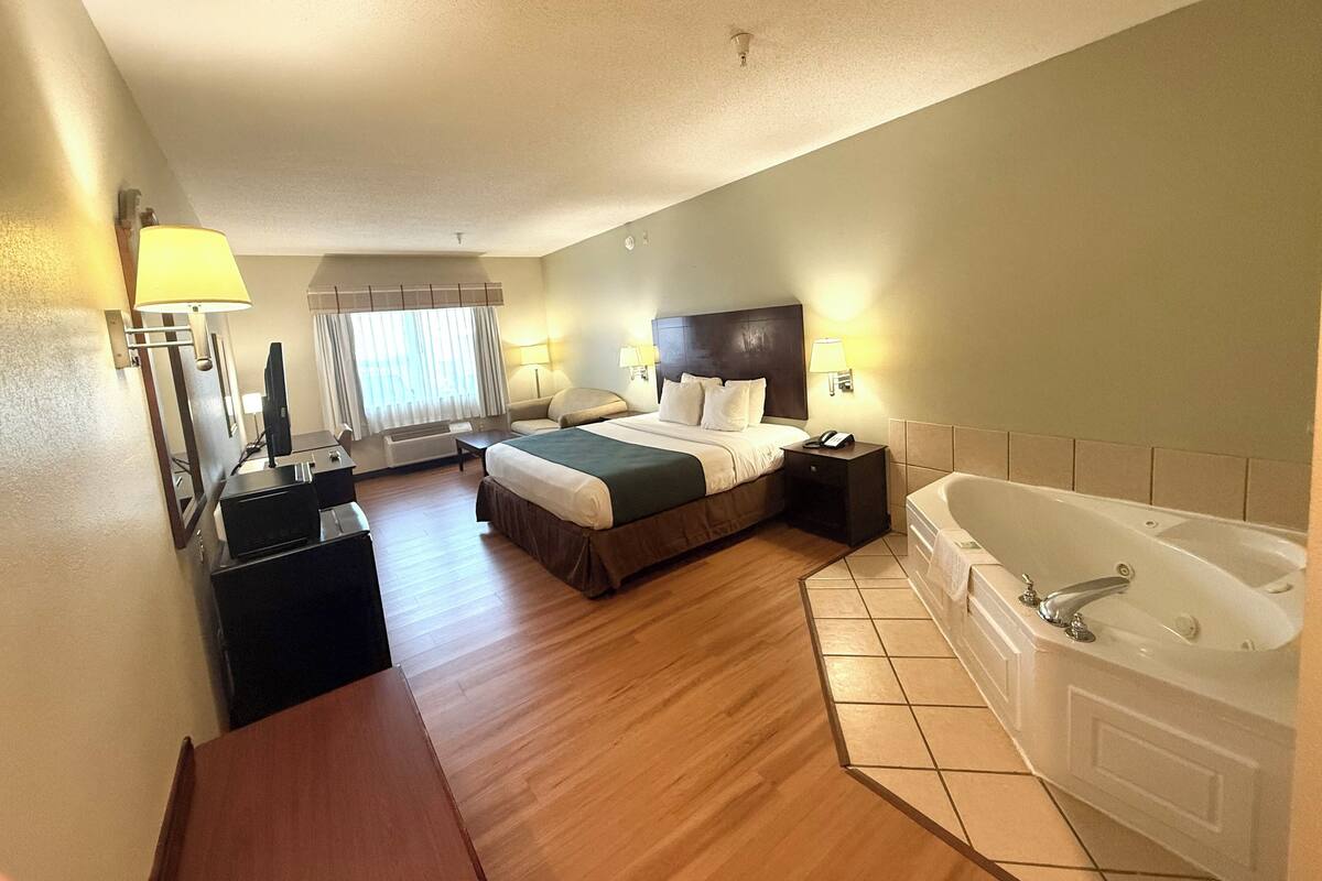 A hotel room with a bed, a desk, a chair, a desk lamp, a telephone, a television, a window with curtains, and a bathtub.