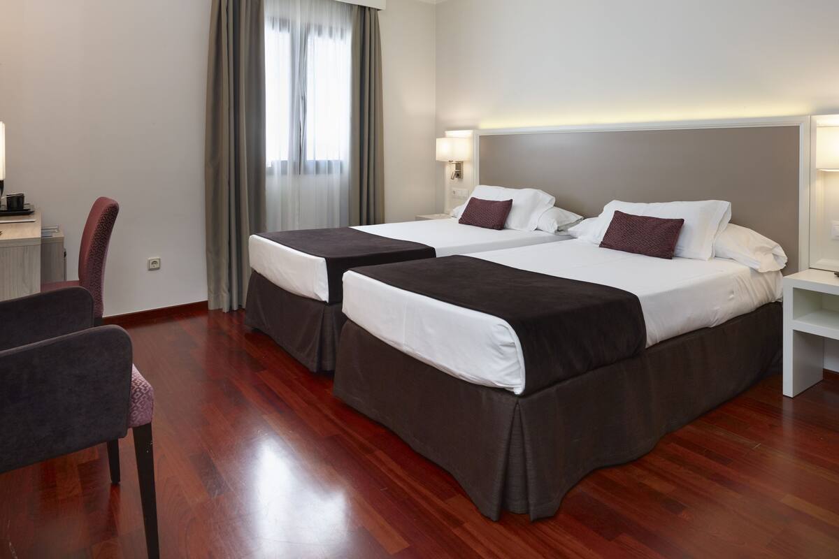 A hotel room with two beds, a desk, a chair, and a window with curtains.