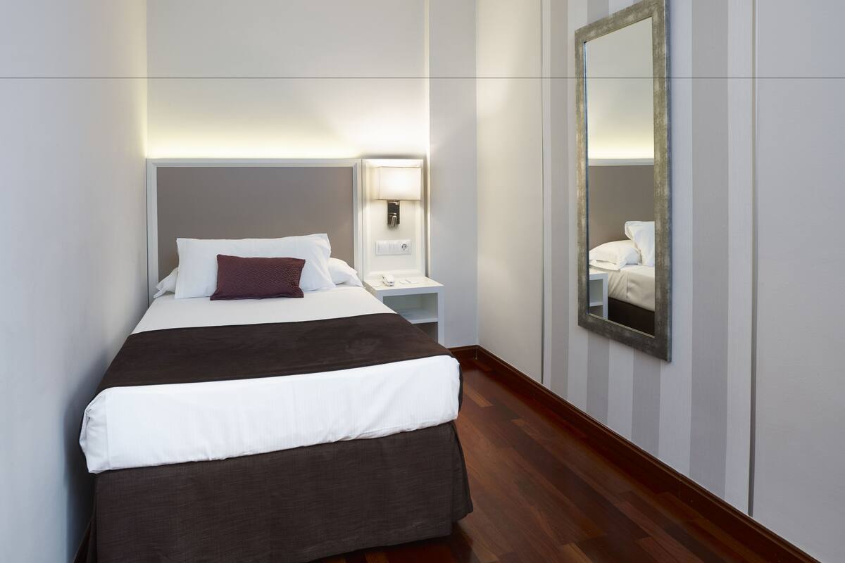 A hotel room with a large bed, a bedside table with a lamp, a mirror, and a wooden floor.