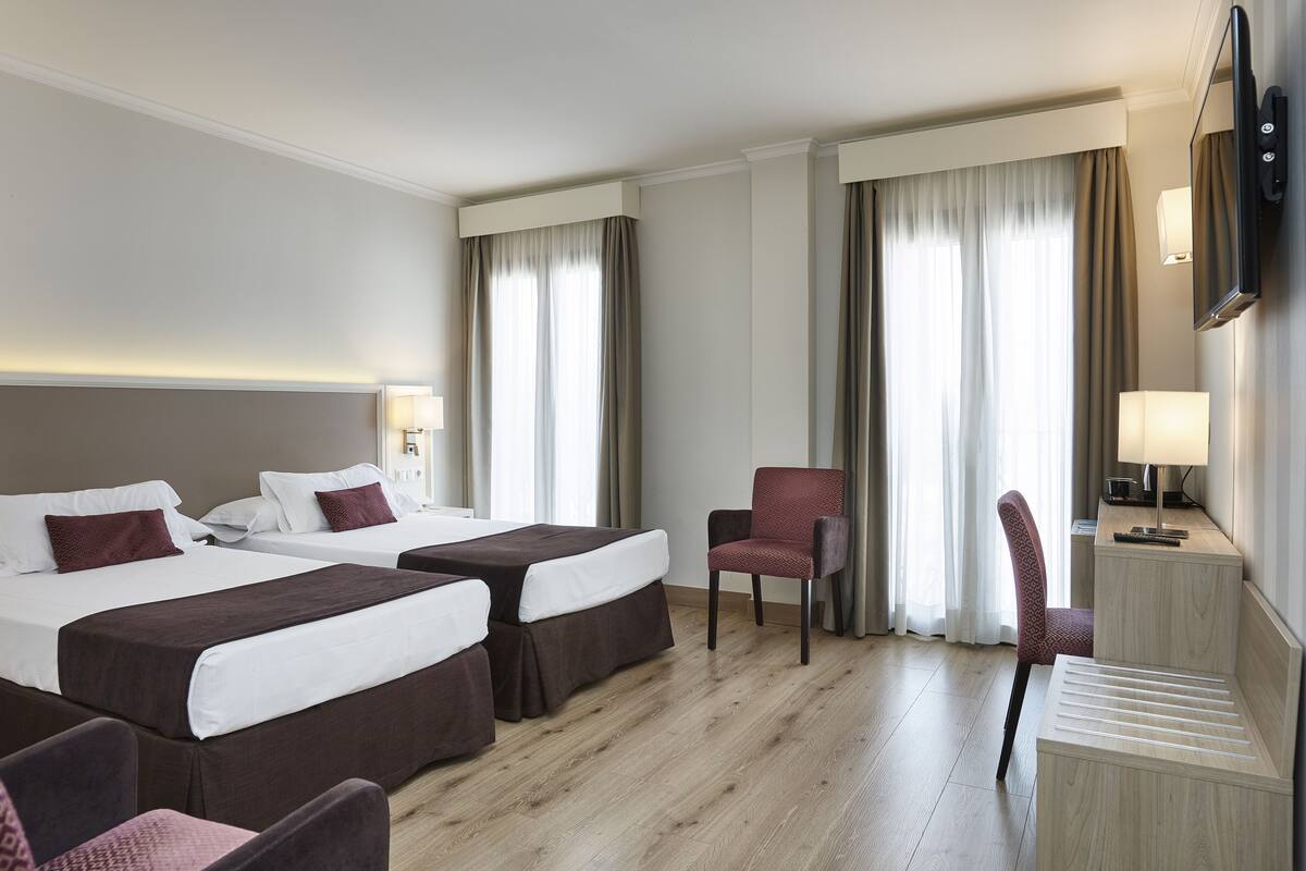 A hotel room with two beds, a desk, chairs, and a TV.