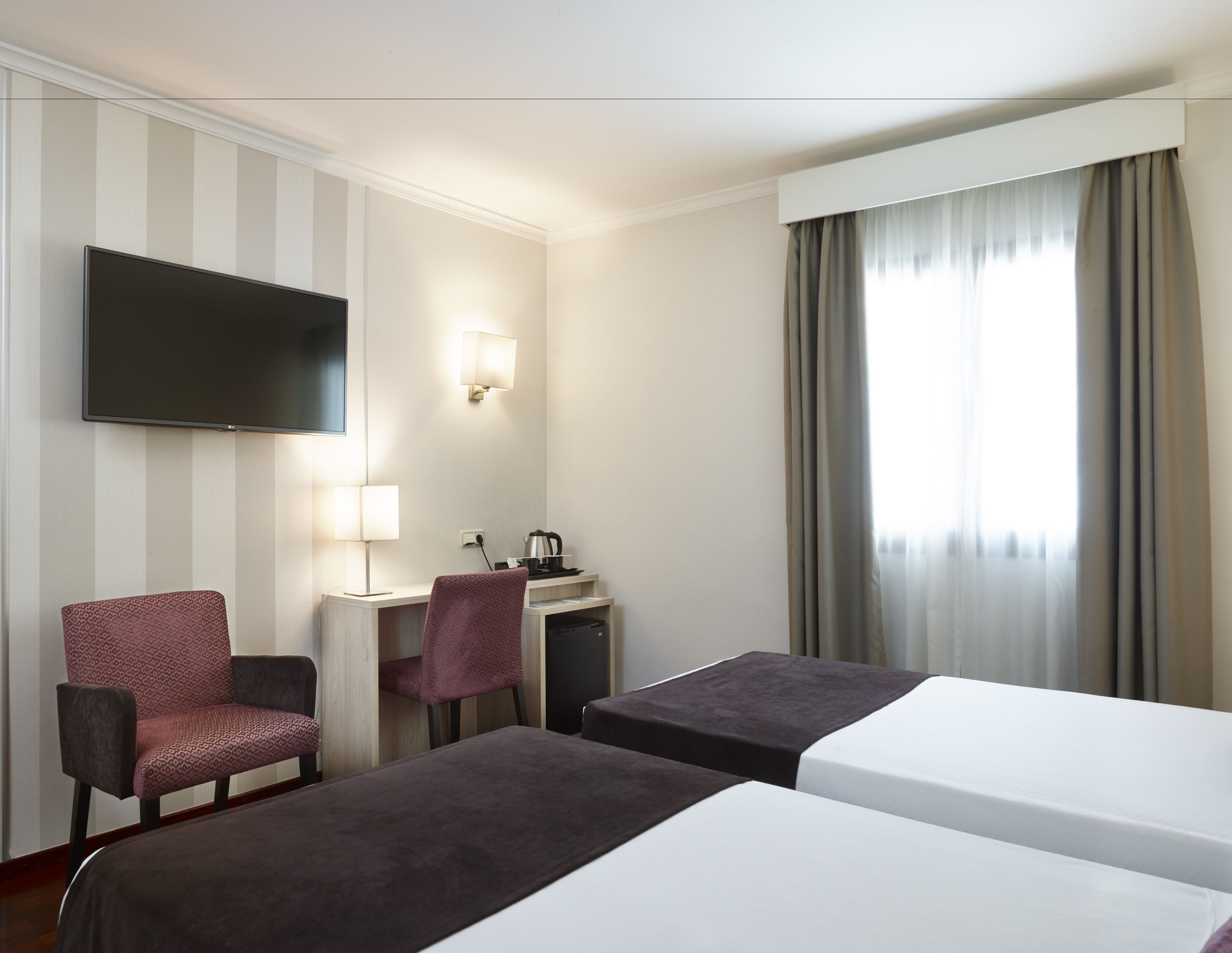 Hotellihuone, jossa on kaksi sänkyä, työpöytä, tuoli, televisio ja ikkuna, jossa on verhot.