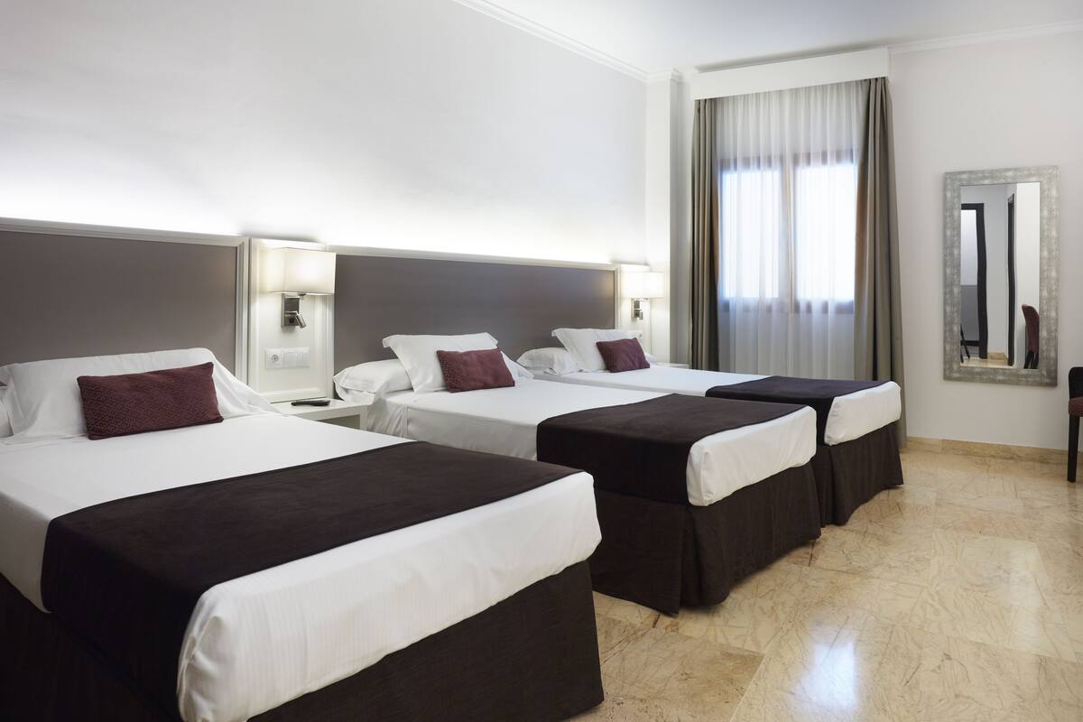 A hotel room with three beds, each with a white and brown bedspread, and a large window with curtains.
