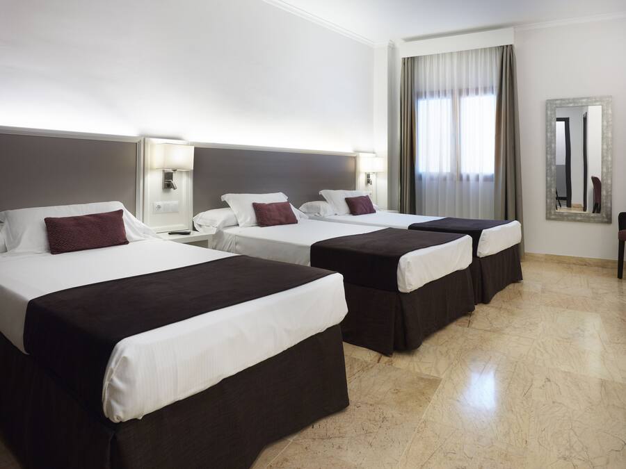 A hotel room with three beds, each with a white and dark-colored bedspread, a headboard, and a nightstand with a lamp.