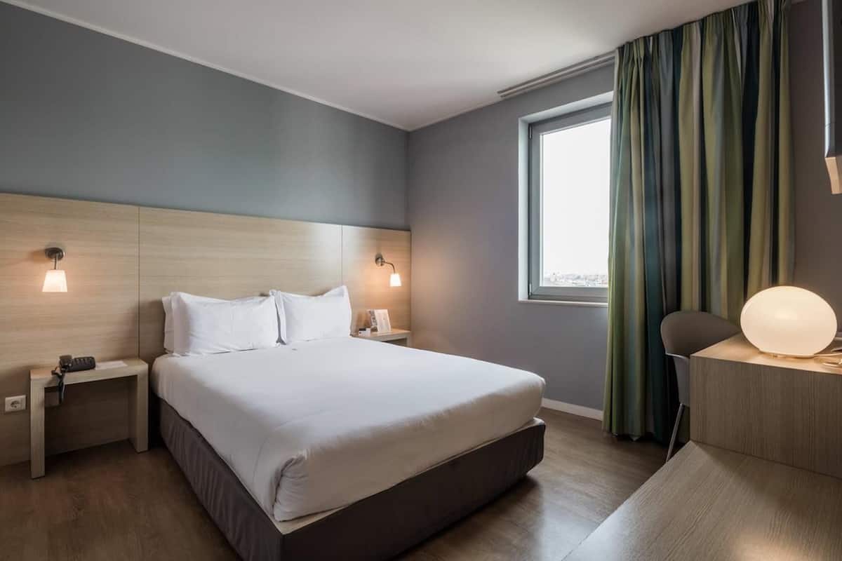 A modern hotel room with a large bed, bedside lamps, a desk with a lamp, and a view of the city through a window.