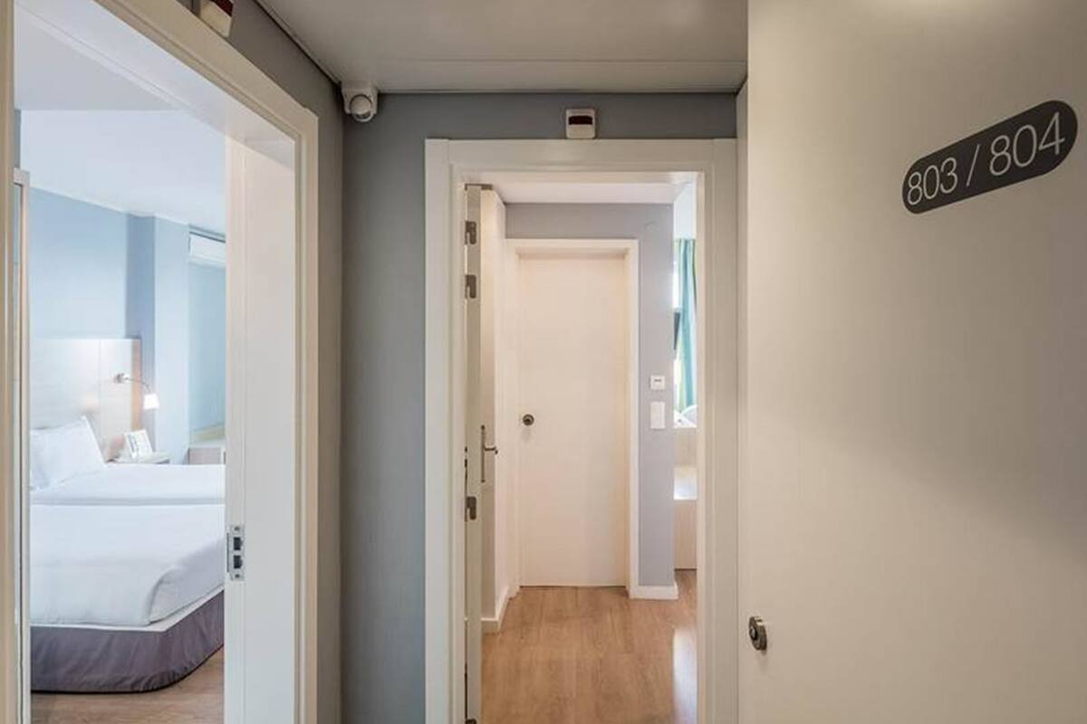 A hotel hallway with a bed visible on the left, a door with a number sign on the right, and a light switch on the wall.