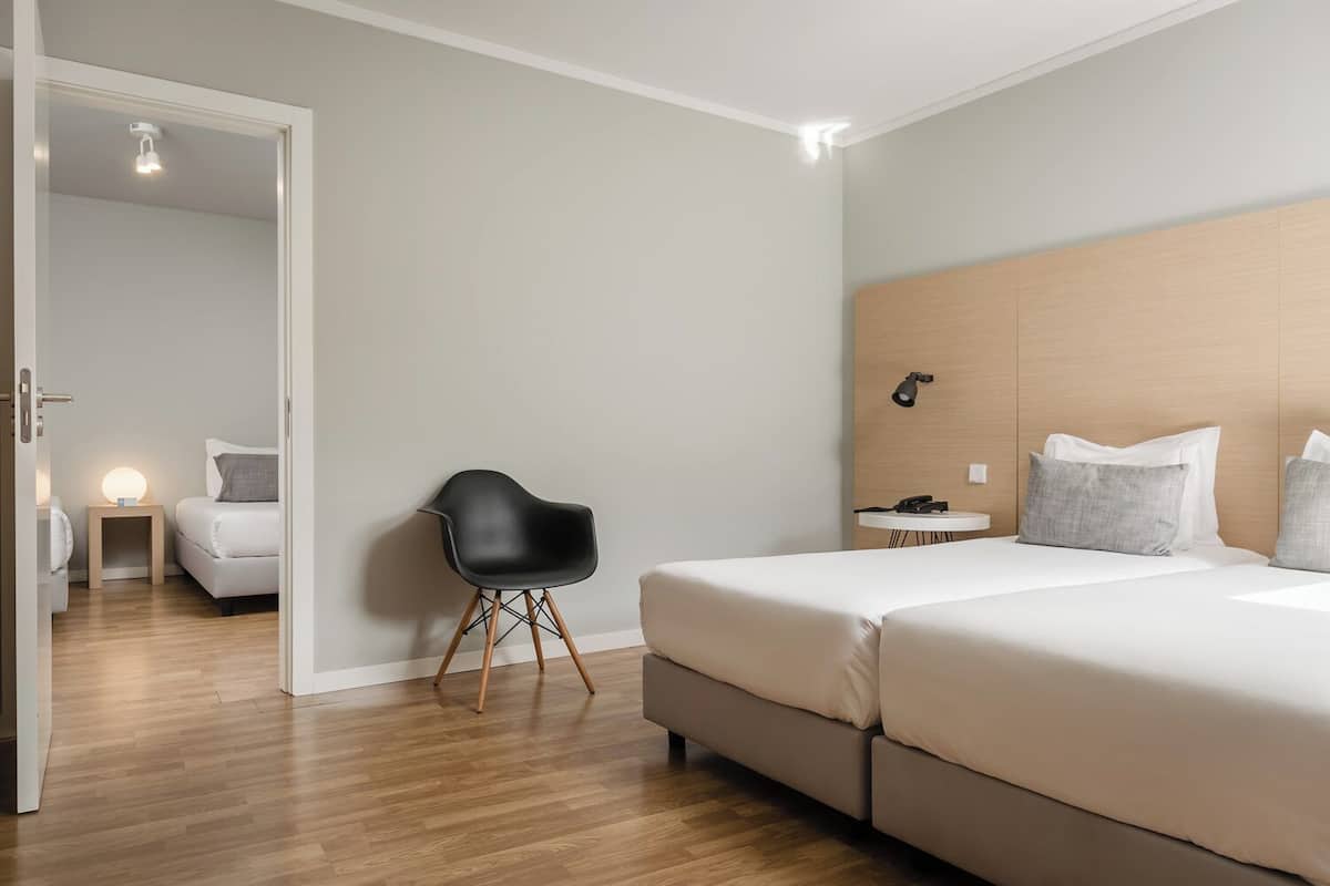 A modern hotel room with a large bed, a small desk, a chair, and a bedside lamp.