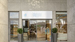 Entrada de hotel con diseño moderno, que presenta un logotipo grande, un cartel de bienvenida y dos plantas en macetas.