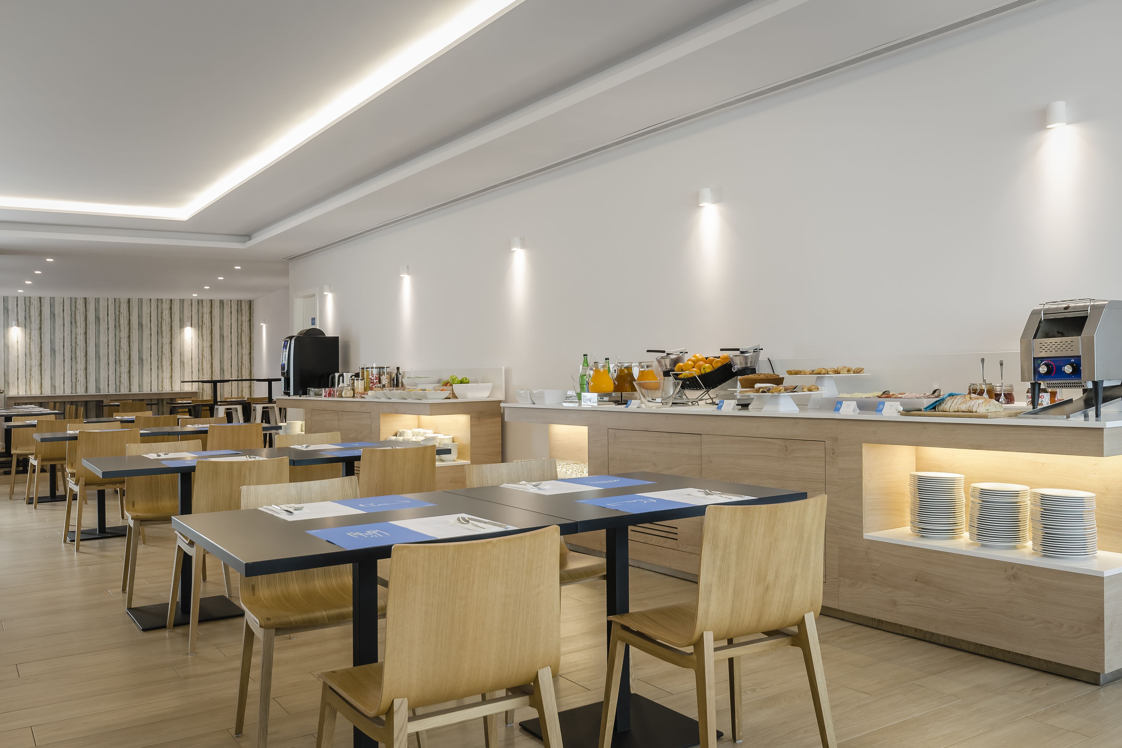 A modern dining area with wooden tables and chairs, a kitchen counter with food items, and a seating area with a striped wall in the background.