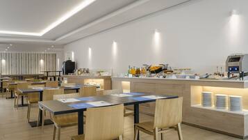 A modern dining area with wooden tables and chairs, a kitchen counter with food items, and a seating area with a striped wall in the background.