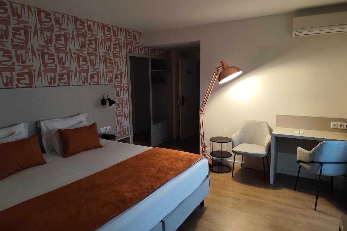 A hotel room with a bed, a desk, and a chair. There is a patterned wall and an air conditioning unit on the wall.