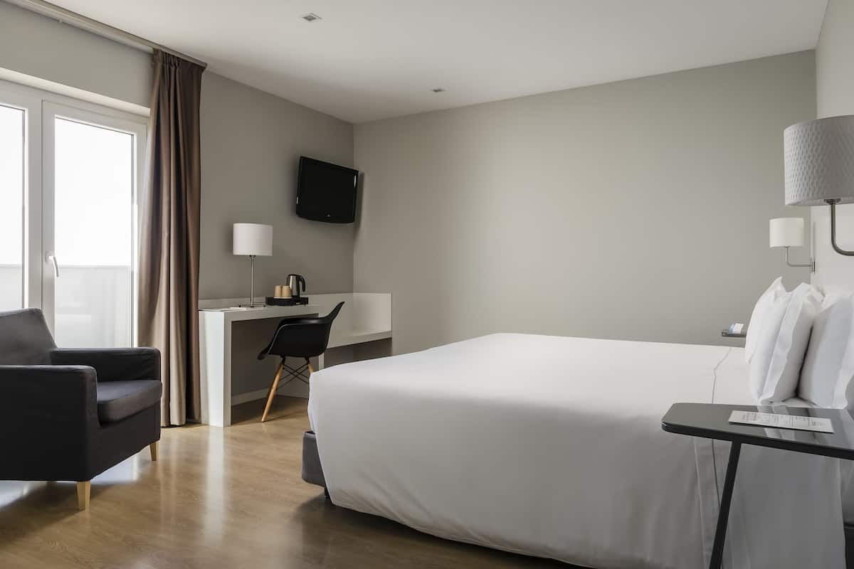 A hotel room with a large bed, a desk with a chair, a TV mounted on the wall, and a window with curtains.