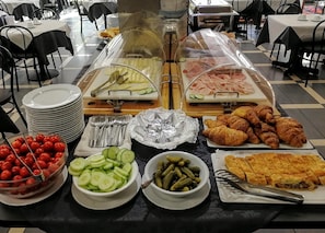 Montaje de bufé con diferentes alimentos, entre ellos pepinos en rodajas, tomates cherry, pepinillos, croissants y una variedad de carnes.