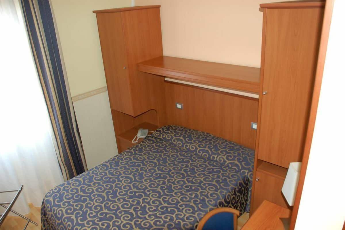 A compact bedroom with a single bed, wooden wardrobe, and a small shelf.