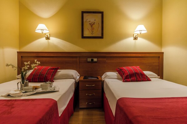 Quarto de hotel com duas camas, cada uma com almofada vermelha e roupa de cama branca. Há mesas de cabeceira com abajures, uma obra de arte emoldurada na parede e uma bandeja com itens de café da manhã na cama da esquerda.