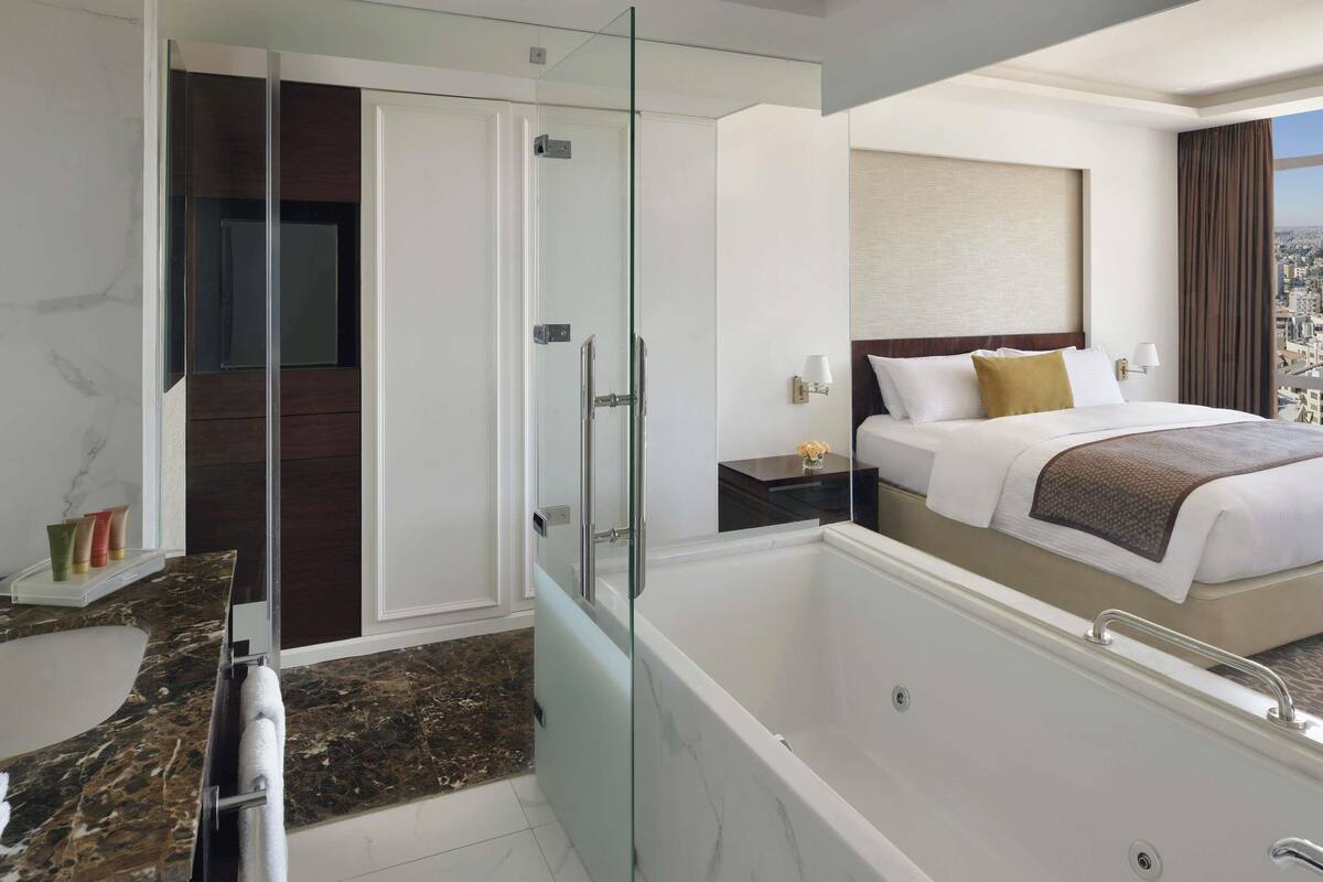 Quarto de hotel moderno com closet envidraçado, cama grande com vista para a cidade, banheiro separado com banheira e piso de mármore.