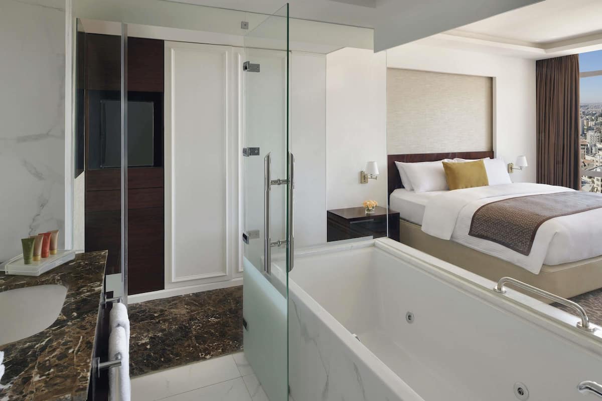 A modern hotel room with a glass-enclosed walk-in closet, a large bed with a city view, a separate bathroom with a bathtub, and a marble-tiled floor.