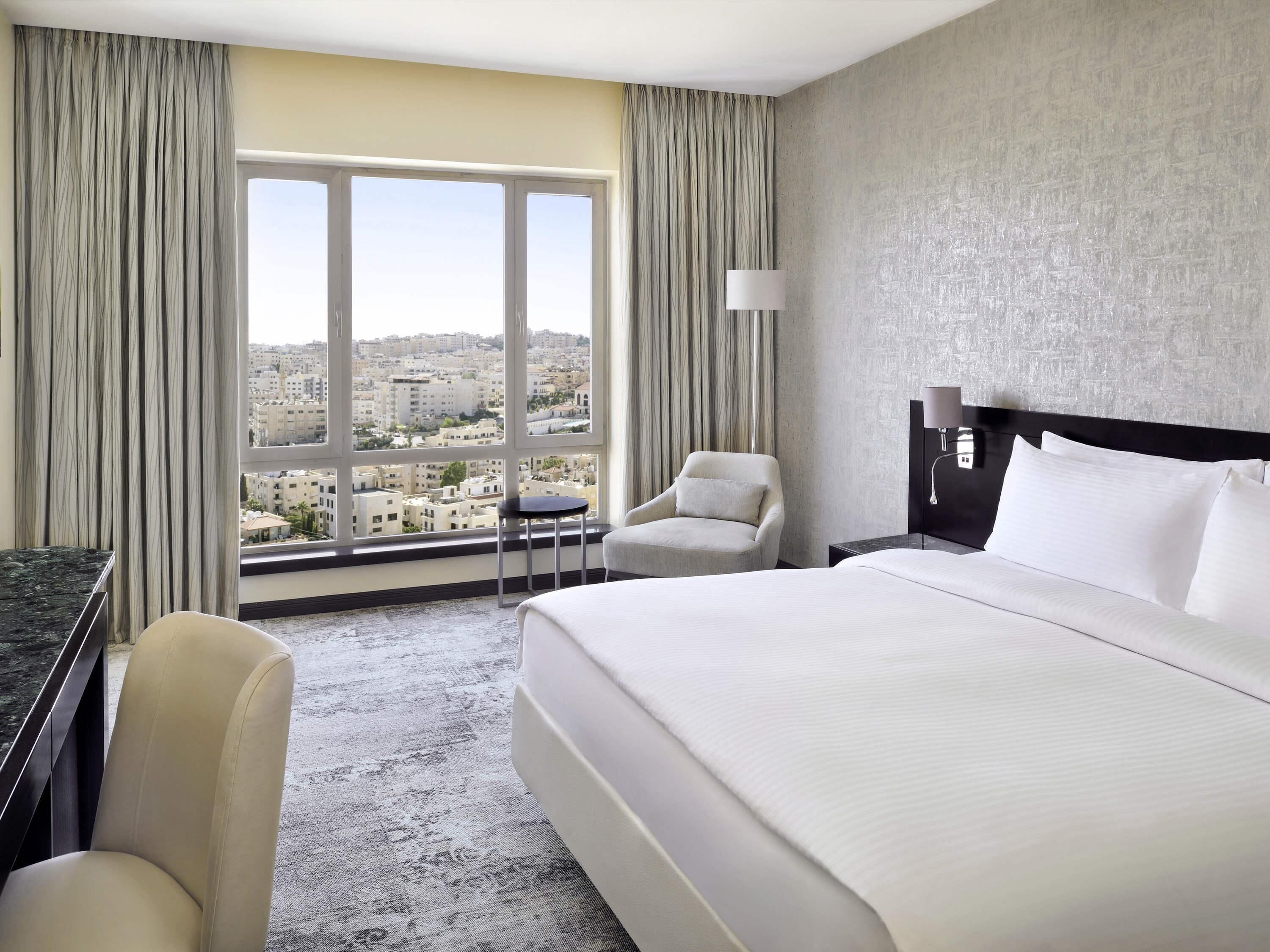 Ein Hotelzimmer mit einem großen Bett, einem Sessel, einem Schreibtisch und Blick auf die Stadt durch große Fenster.