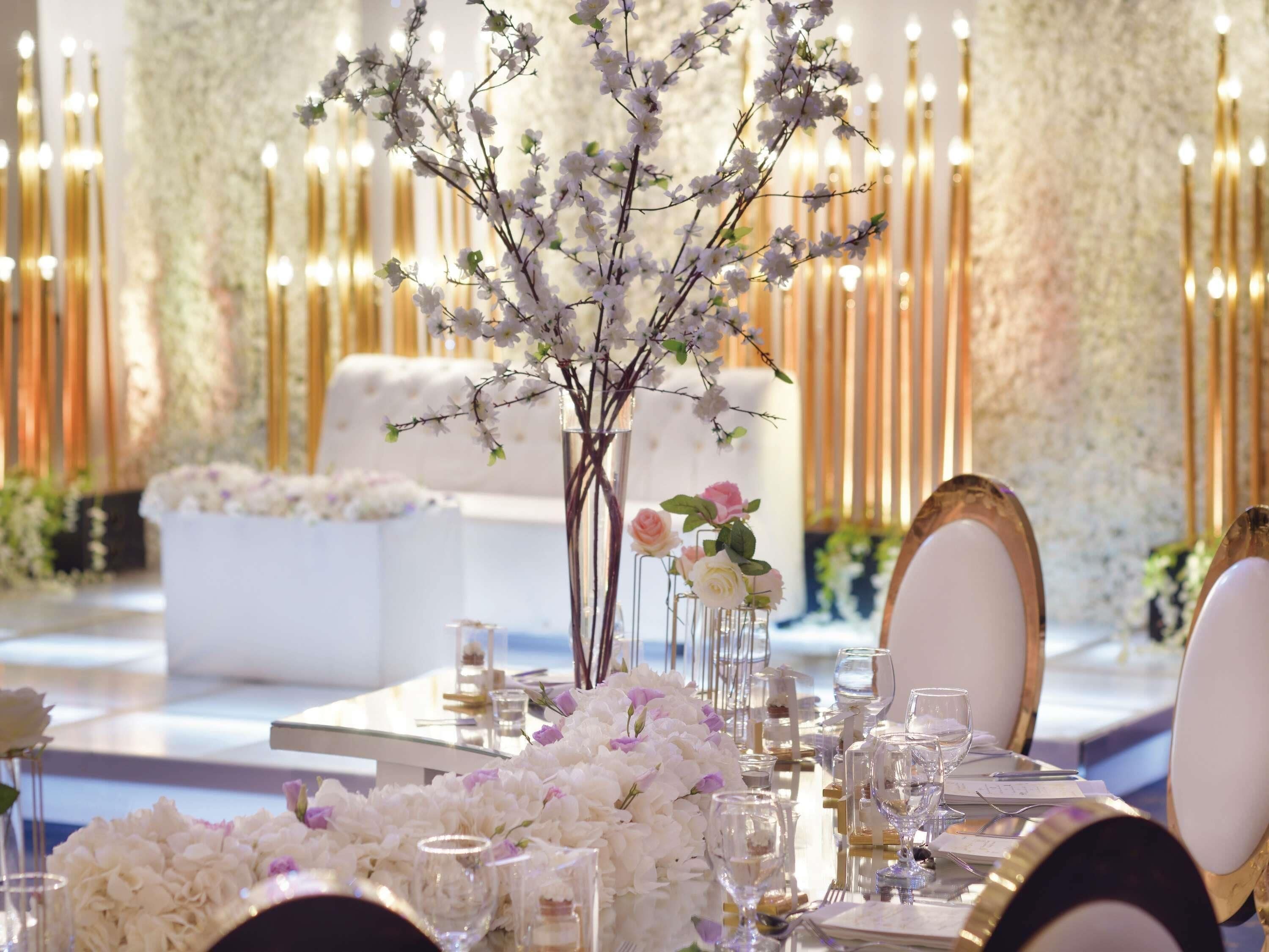 A formal dining table set with glassware, plates, and cutlery, surrounded by white floral arrangements and a tall vase with branches.