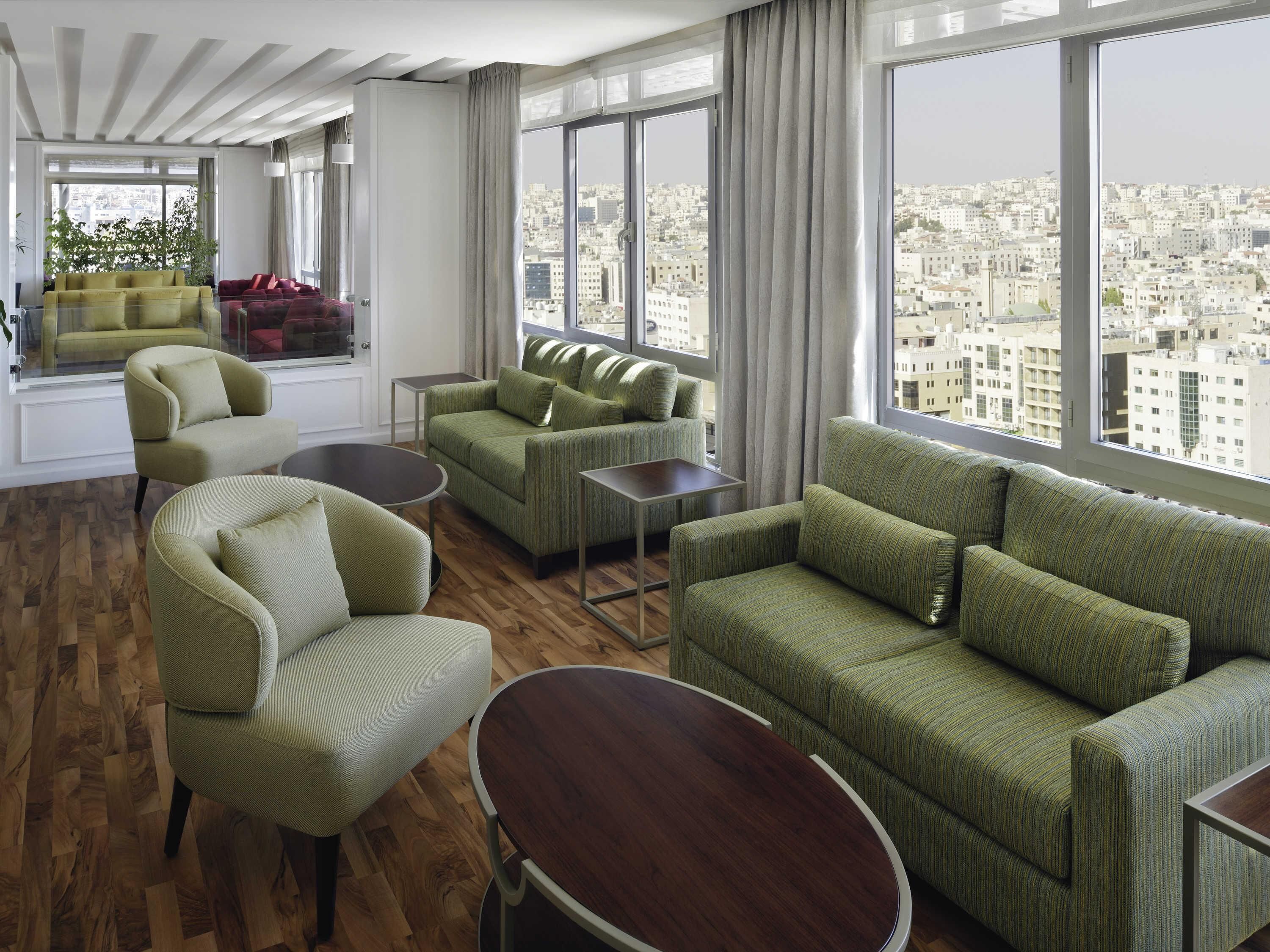 A modern living room with green sofas, a round wooden table, and large windows offering a city view.