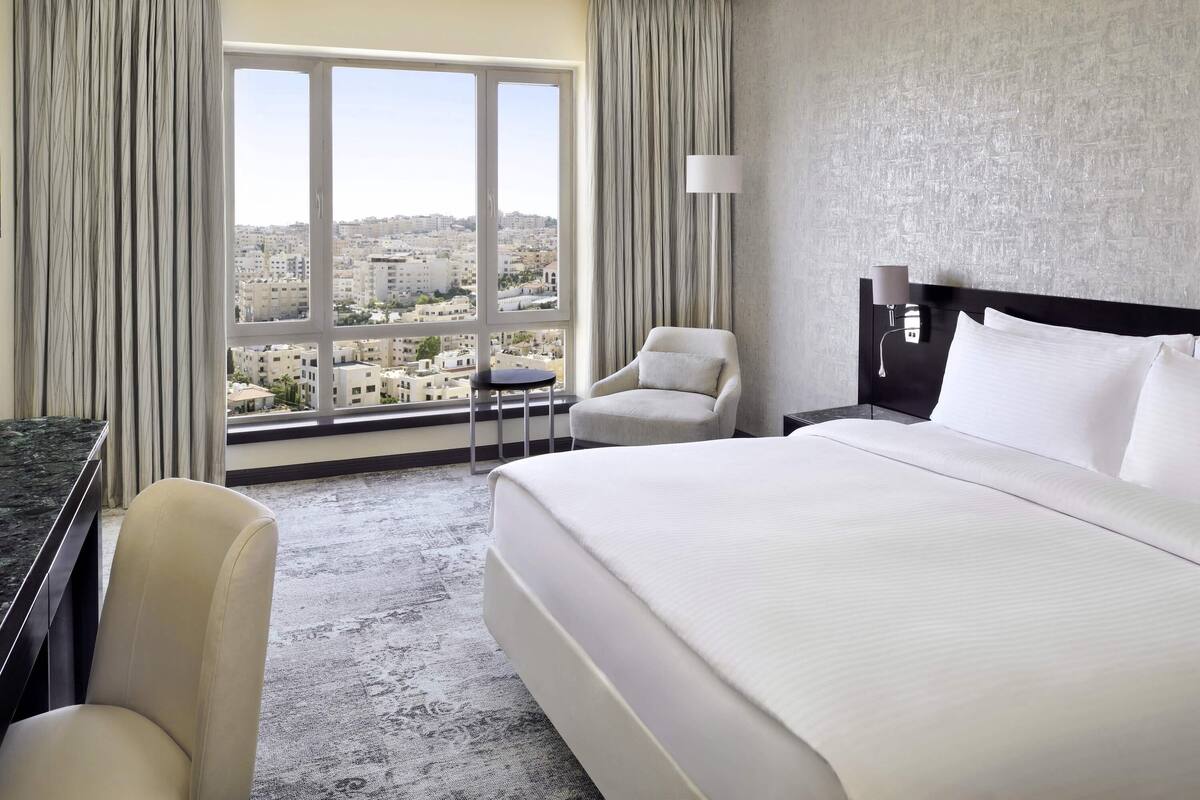 Habitación de hotel con una cama grande, una silla, un escritorio y vistas de la ciudad a través de amplias ventanas.