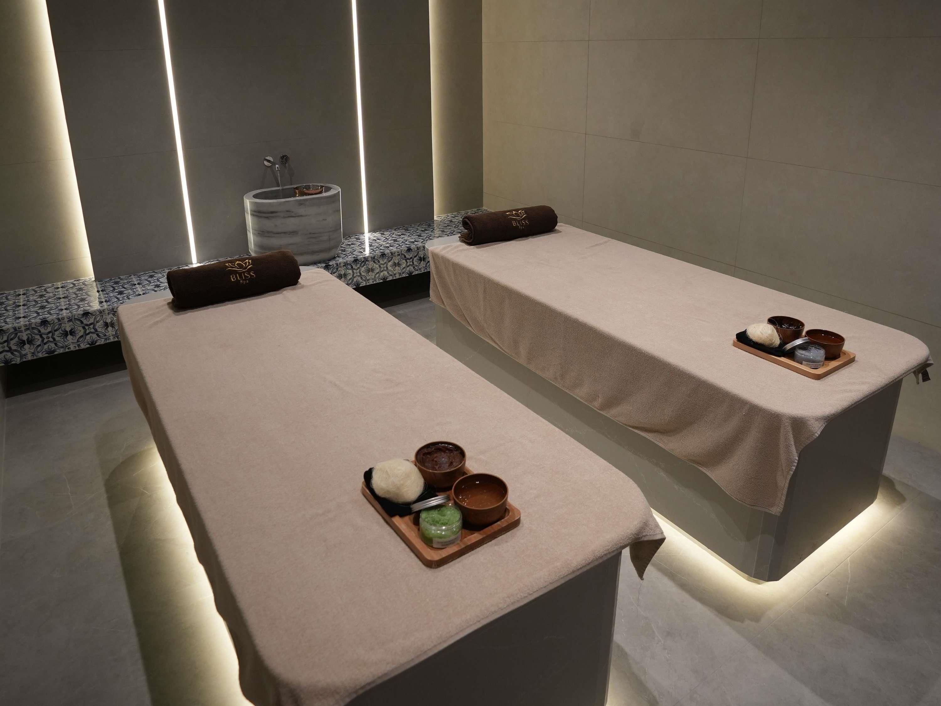 Dos camillas de masaje con toallas y cuencos de comida, un lavabo de mármol y un banco con motivos decorativos en un entorno de spa.