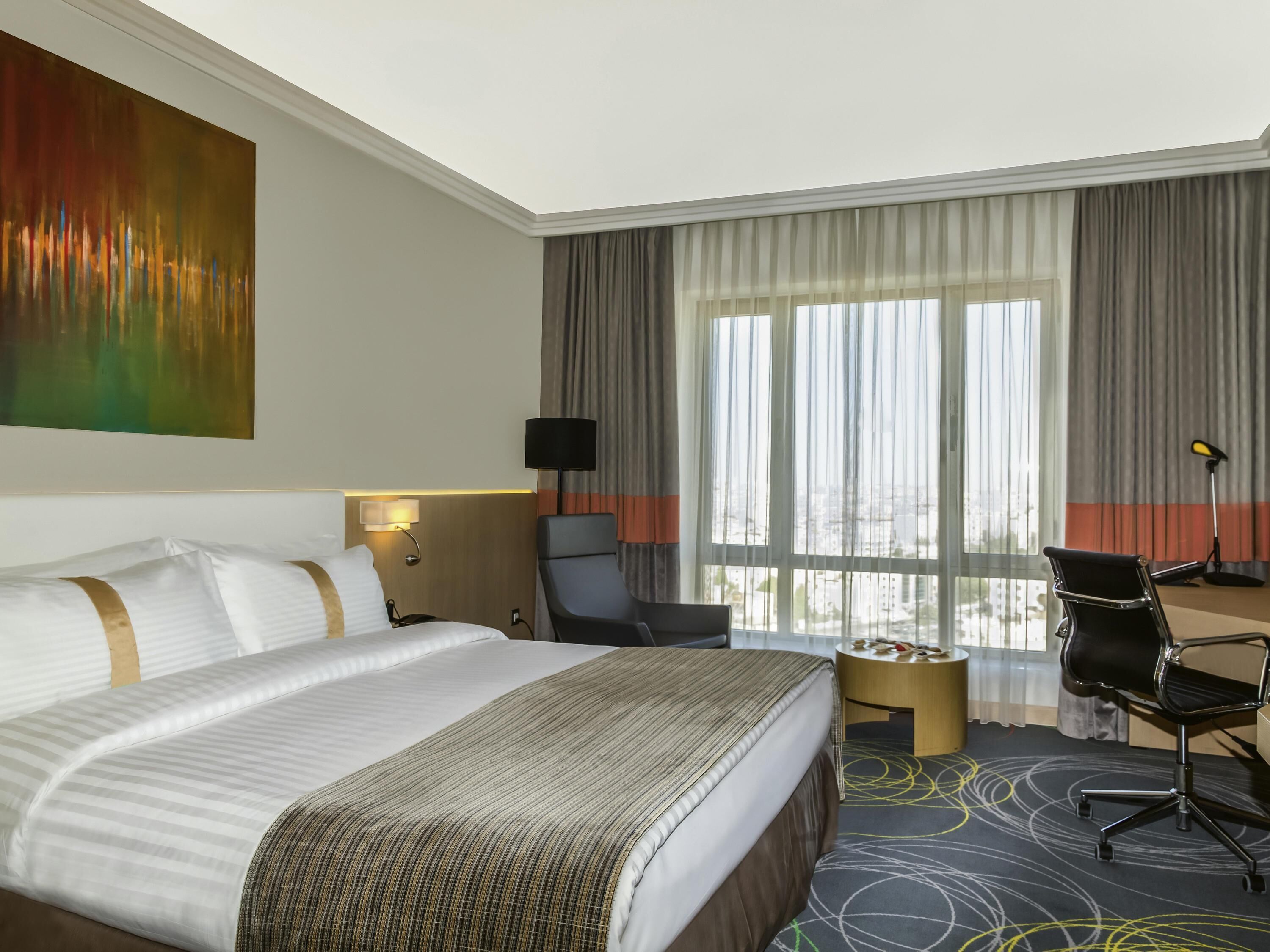 Ein Hotelzimmer mit einem großen Bett, einem Schreibtisch, einem Sessel und einer Lampe, mit Blick auf die Stadt durch große Fenster.