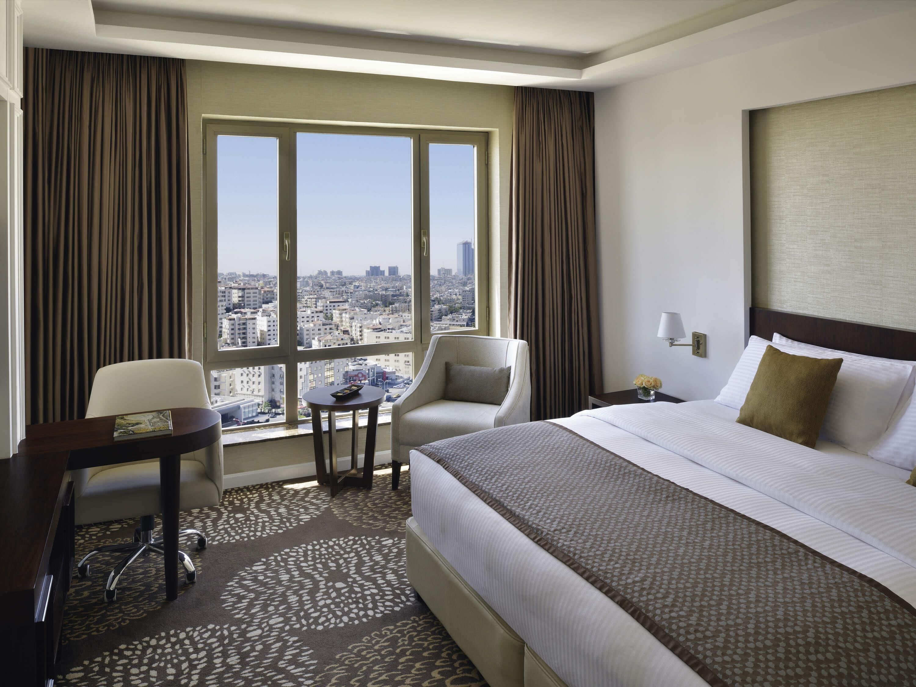 Een hotelkamer met een groot bed, een bureau, een stoel en uitzicht op de stad door grote ramen.