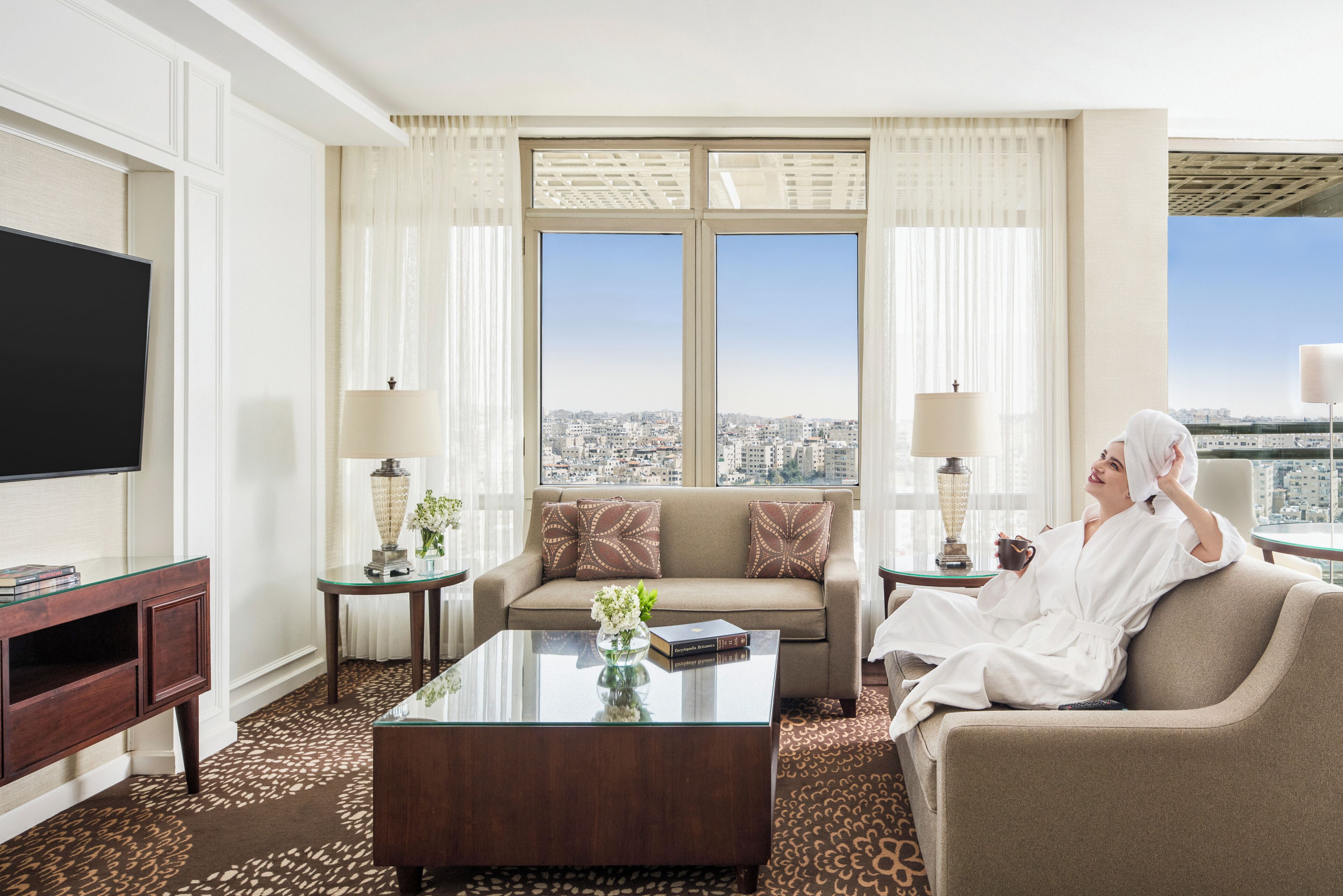 A hotel room with a sofa, a coffee table, a TV, and a view of the city.