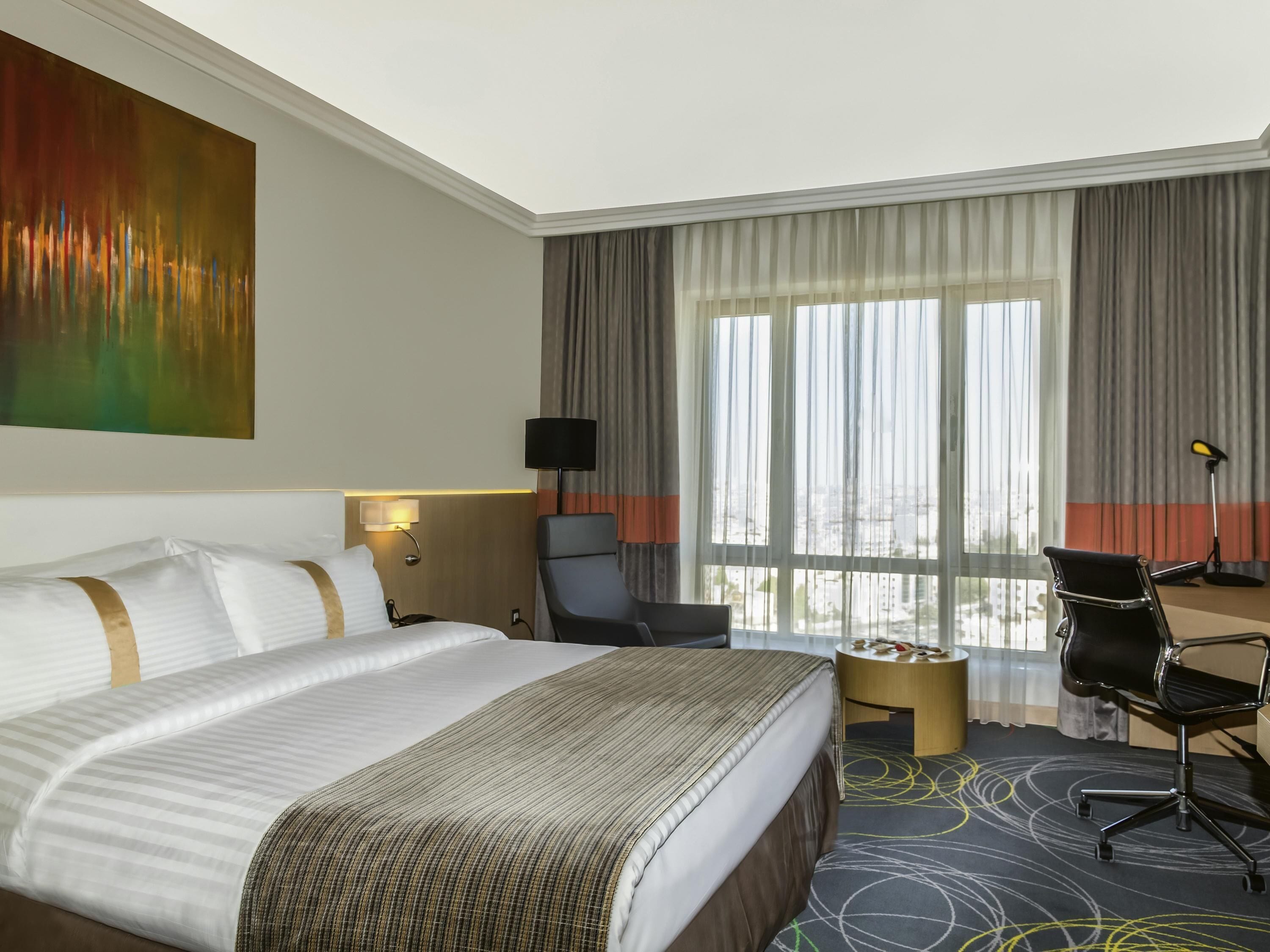 A hotel room with a large bed, a desk, a chair, a lamp, and a view of the city through large windows.