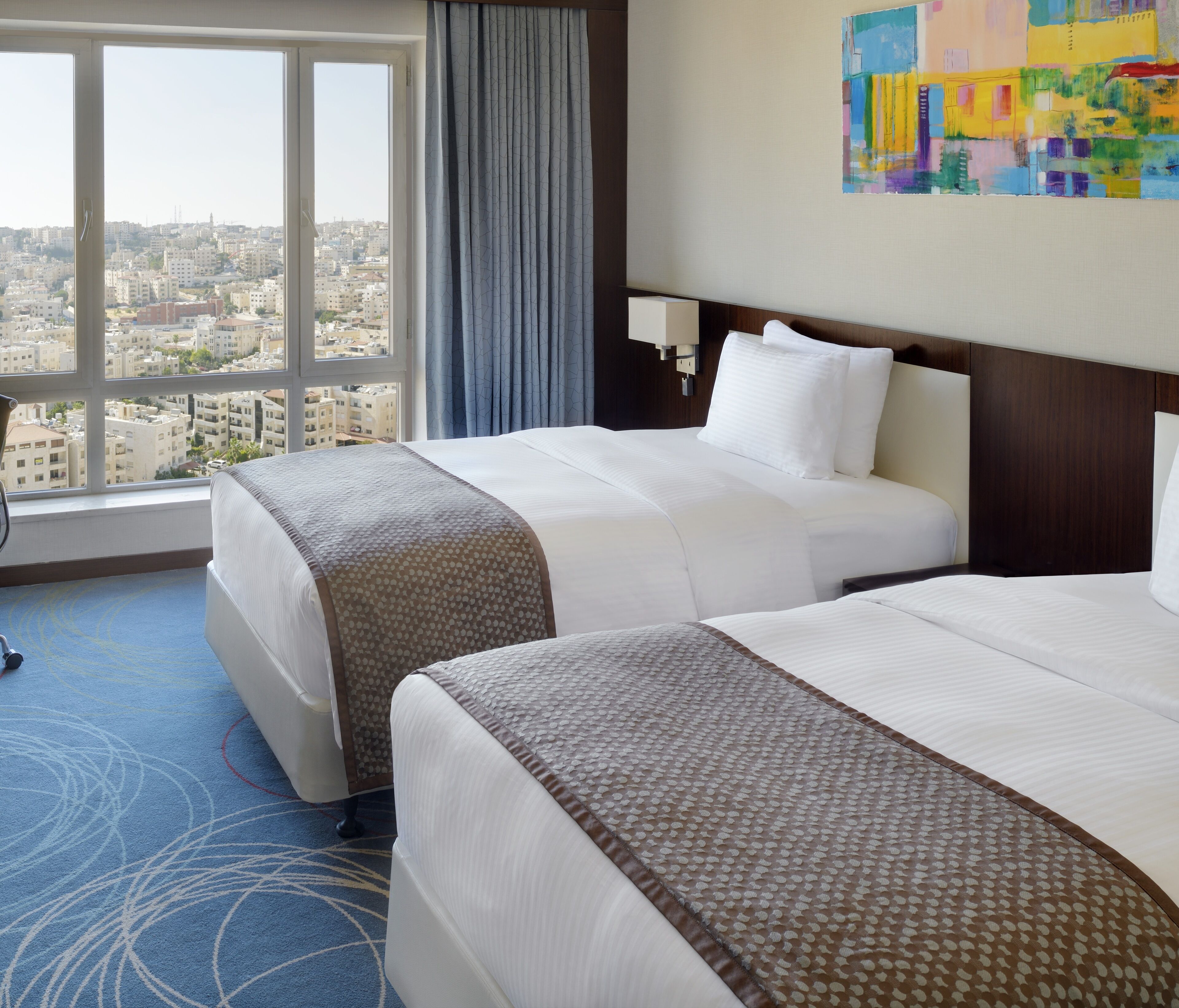 Habitación de hotel con dos camas, un ventanal amplio con vistas a la ciudad, alfombra azul y un cuadro abstracto de colores en la pared.