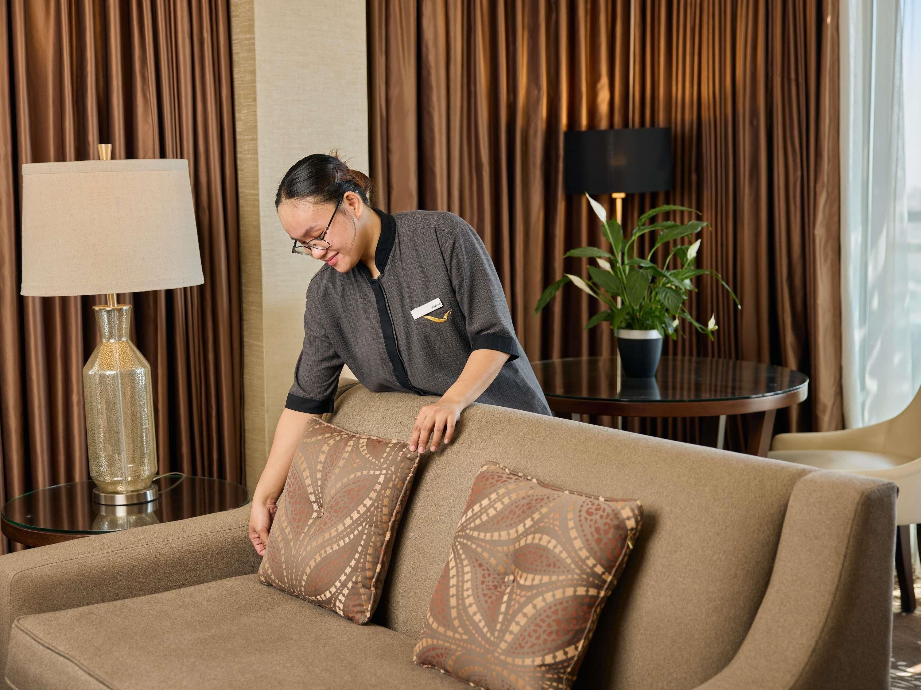 A person adjusting a sofa with patterned cushions in a room with a lamp and a potted plant.