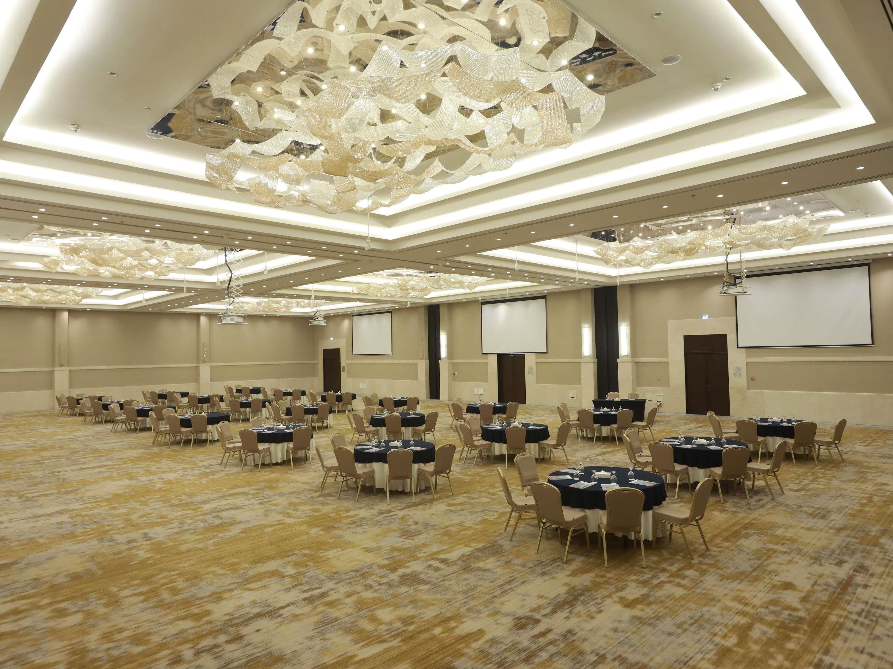 A large conference room with multiple tables and chairs, a projector screen, and a decorative ceiling.