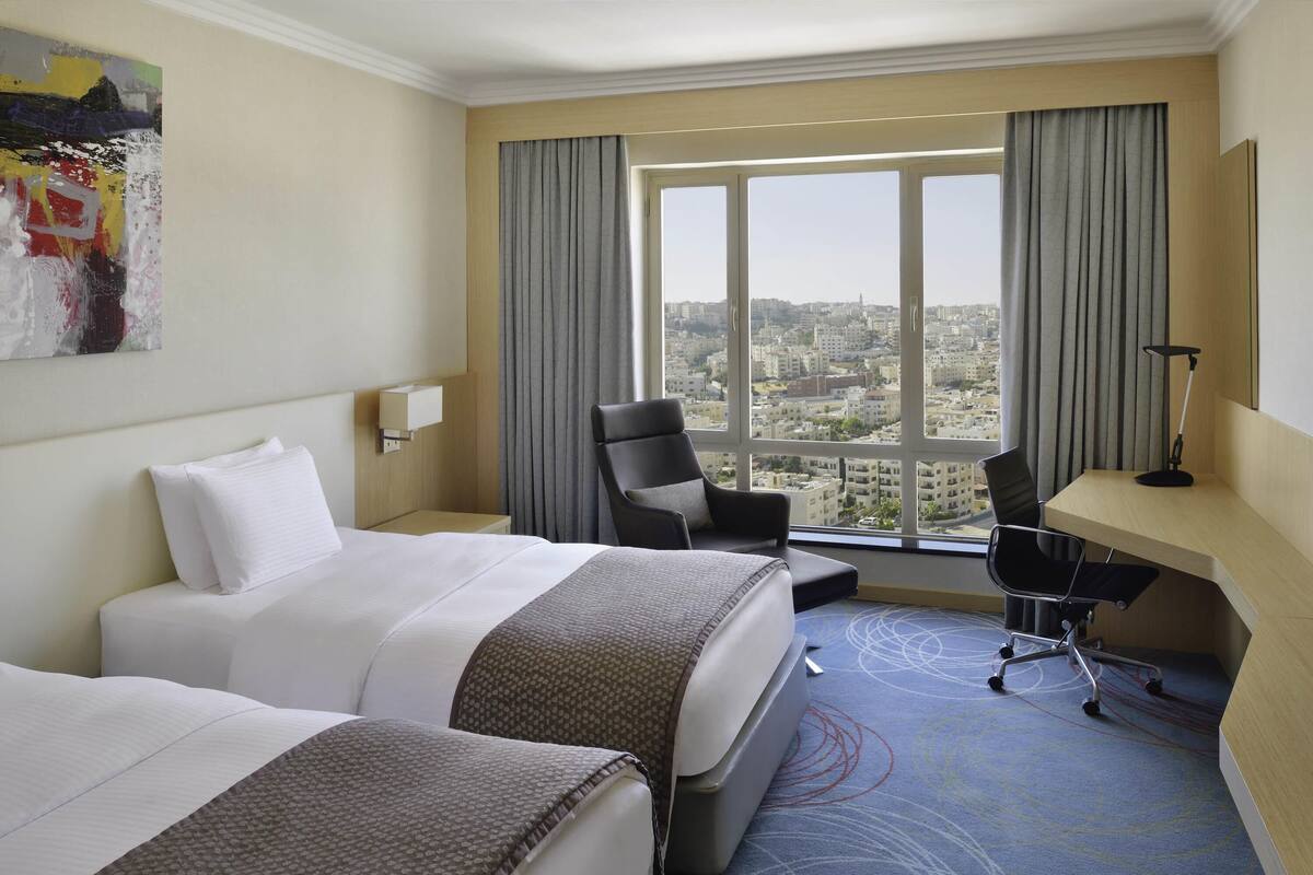 Habitación de hotel con dos camas, un escritorio, una silla, una lámpara y un ventanal con vistas a la ciudad.