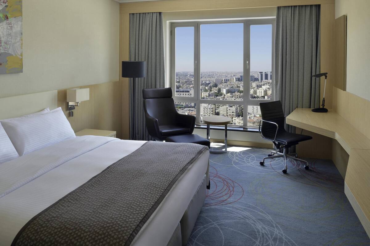 Una habitación de hotel con una cama grande, un escritorio, dos sillas, una mesita, una lámpara y vistas a un paisaje urbano a través de amplios ventanales.