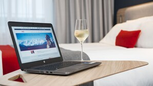 A laptop on a wooden table with a glass of white wine beside it, in a hotel room with a bed, red pillows, and a view of mountains through the window. - Holiday Inn Express Bath by IHG (Bath)