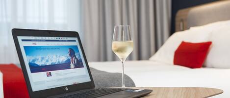 A laptop on a wooden table with a glass of white wine beside it, in a hotel room with a bed, red pillows, and a view of mountains through the window.