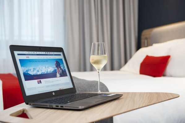 Un ordinateur portable est posé sur une table en bois, à côté se trouve un verre de vin blanc. La scène se déroule dans une chambre d’hôtel, où l’on aperçoit un lit, des oreillers rouges et, par la fenêtre, une vue sur les montagnes.
