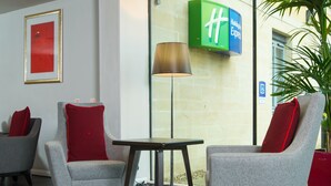 A lobby with two grey armchairs with red cushions, a small round table, and a floor lamp. A green sign with a white 'H' is visible on the wall. - Holiday Inn Express Bath by IHG (Bath)