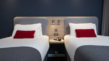 Een hotelkamer met twee bedden, elk voorzien van een grijs hoofdeinde en witte beddengoed, met rode kussens. Tussen de bedden staat een houten nachtkastje met een telefoon en een kleine lamp.