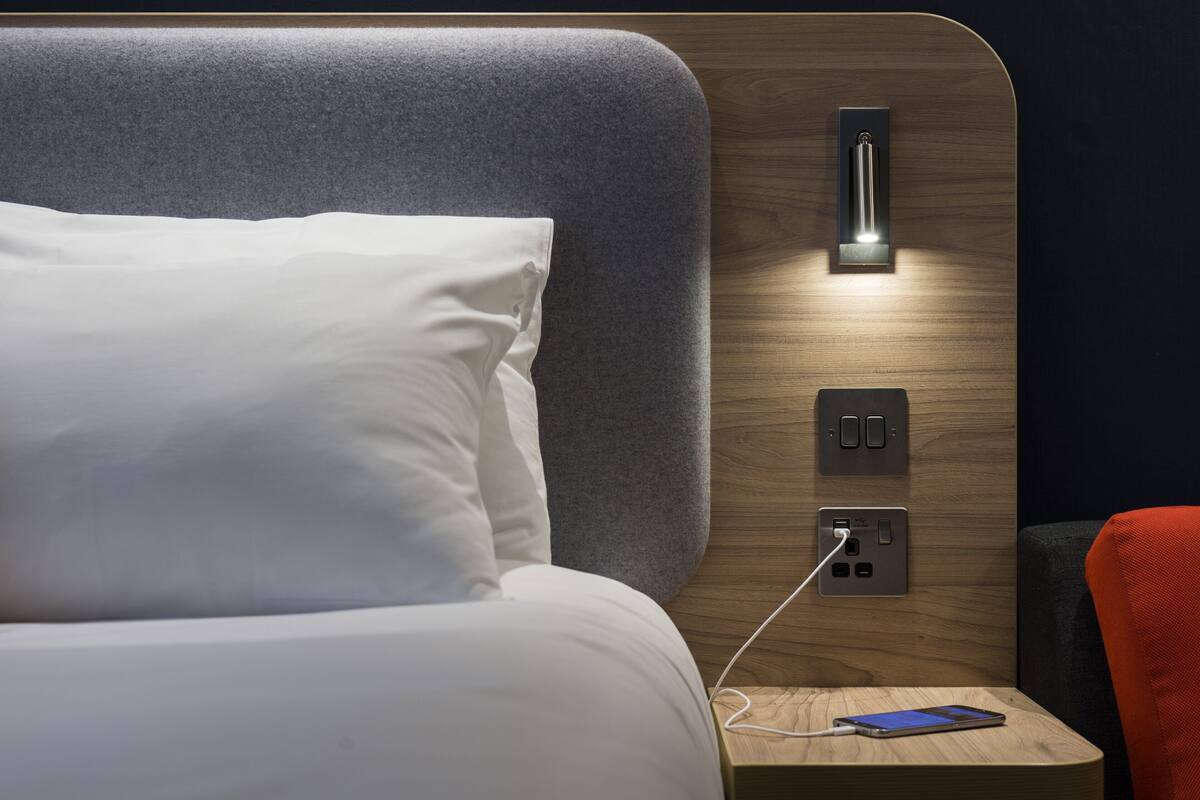 A modern hotel room with a neatly made bed, a bedside table with a smartphone charging, and a wall-mounted light fixture.