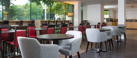 Un restaurante moderno con mesas y sillas redondas, amplias ventanas que dan a vistas de árboles, y una decoración interior limpia y contemporánea.
