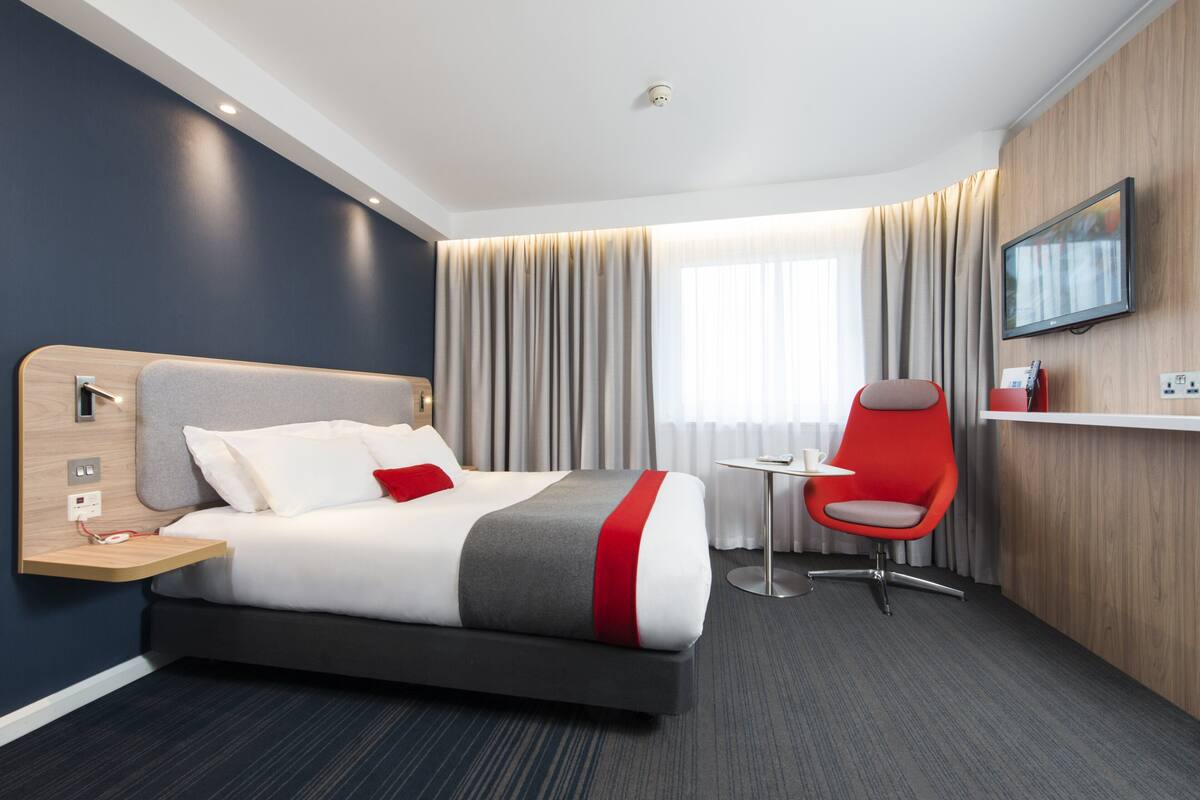 A modern hotel room with a bed, a desk, a chair, a TV, and a window with curtains.