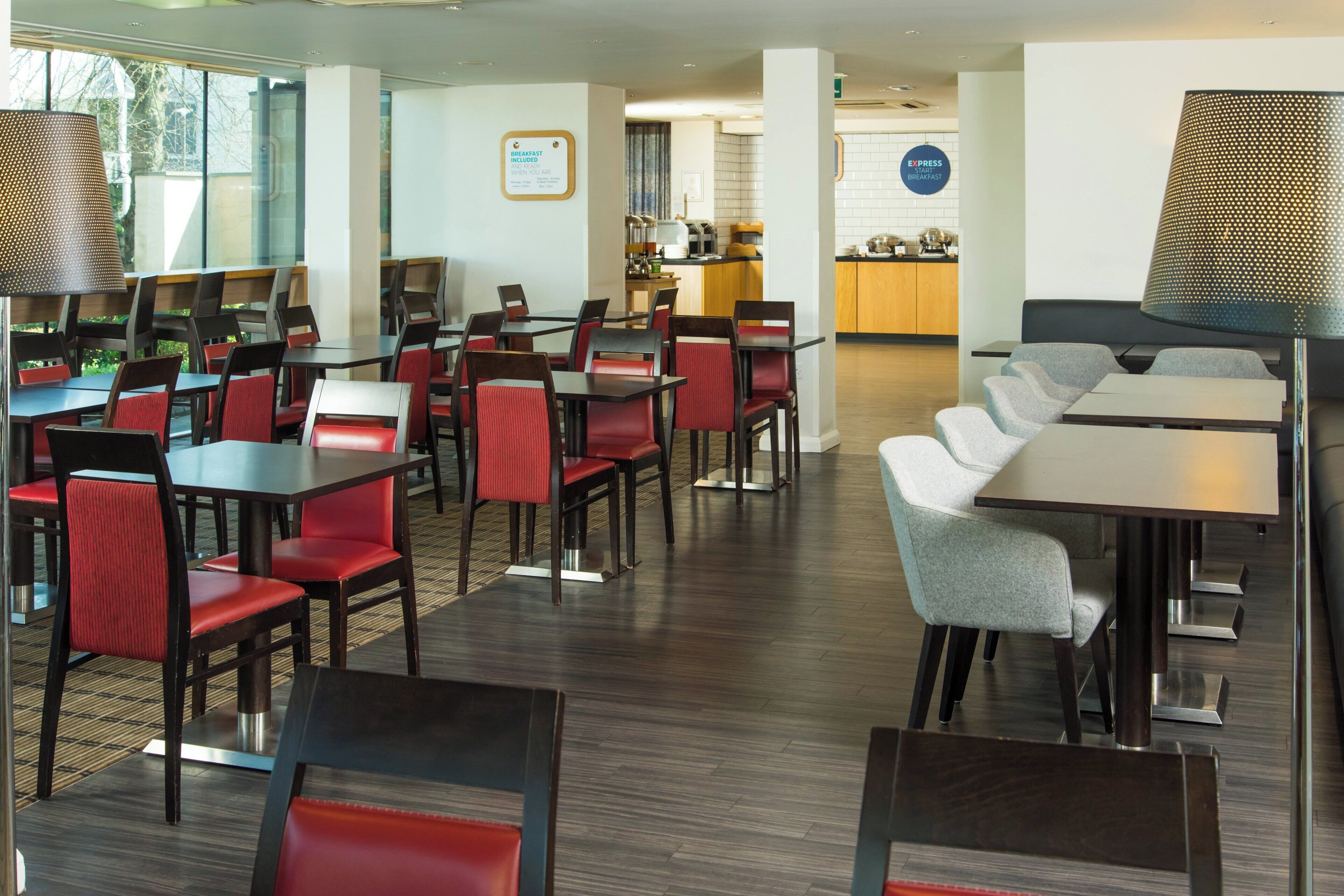 A hotel dining area with multiple tables and chairs, a kitchen in the background, and a sign indicating breakfast is included.