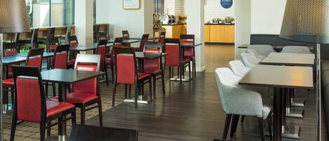 A hotel dining area with multiple tables and chairs, a kitchen in the background, and a sign indicating breakfast is included.