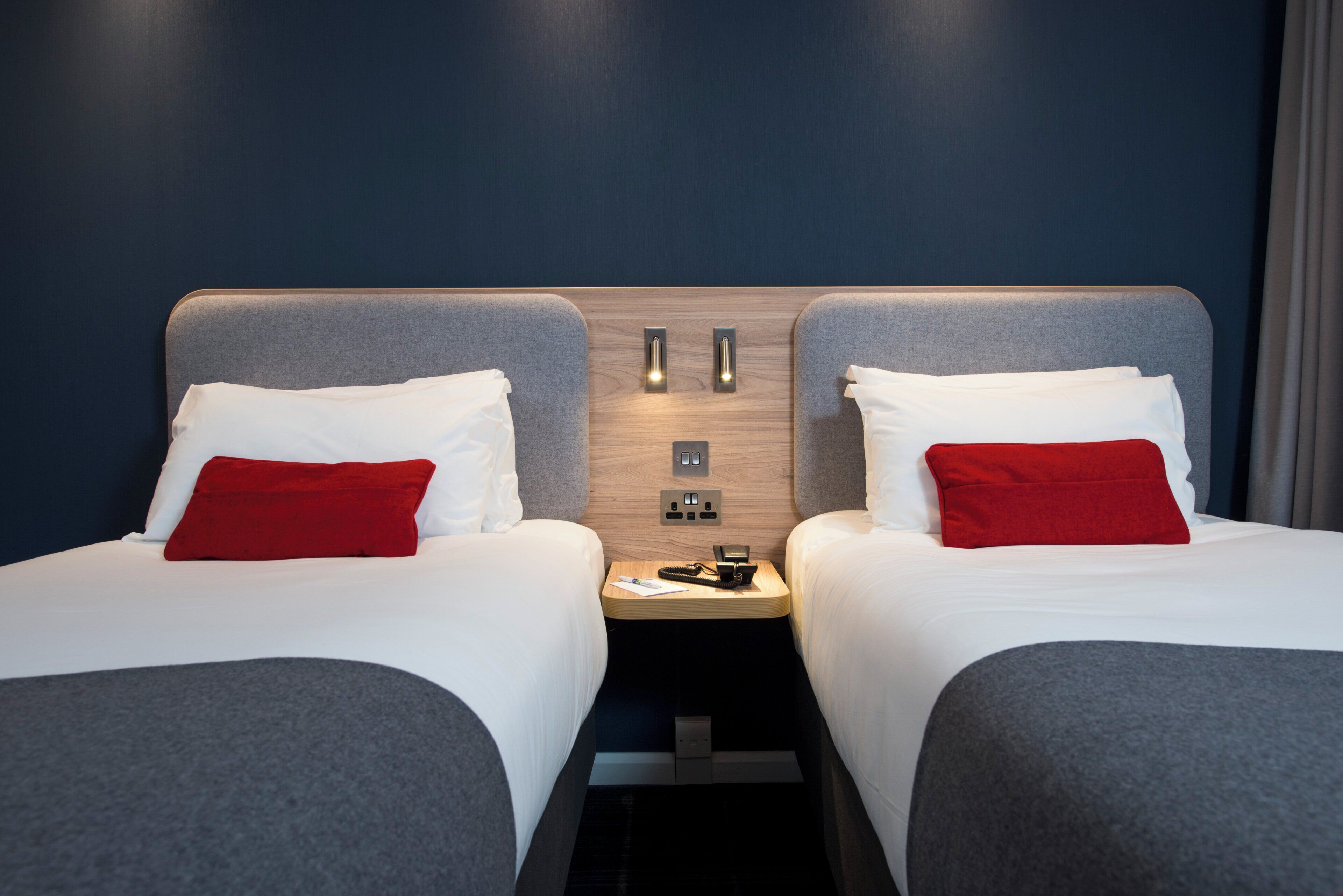 Een hotelkamer met twee bedden, elk met een grijs hoofdeinde, witte beddengoed en een rood kussen. Daartussen staat een houten nachtkastje met een telefoon en een kleine lamp.