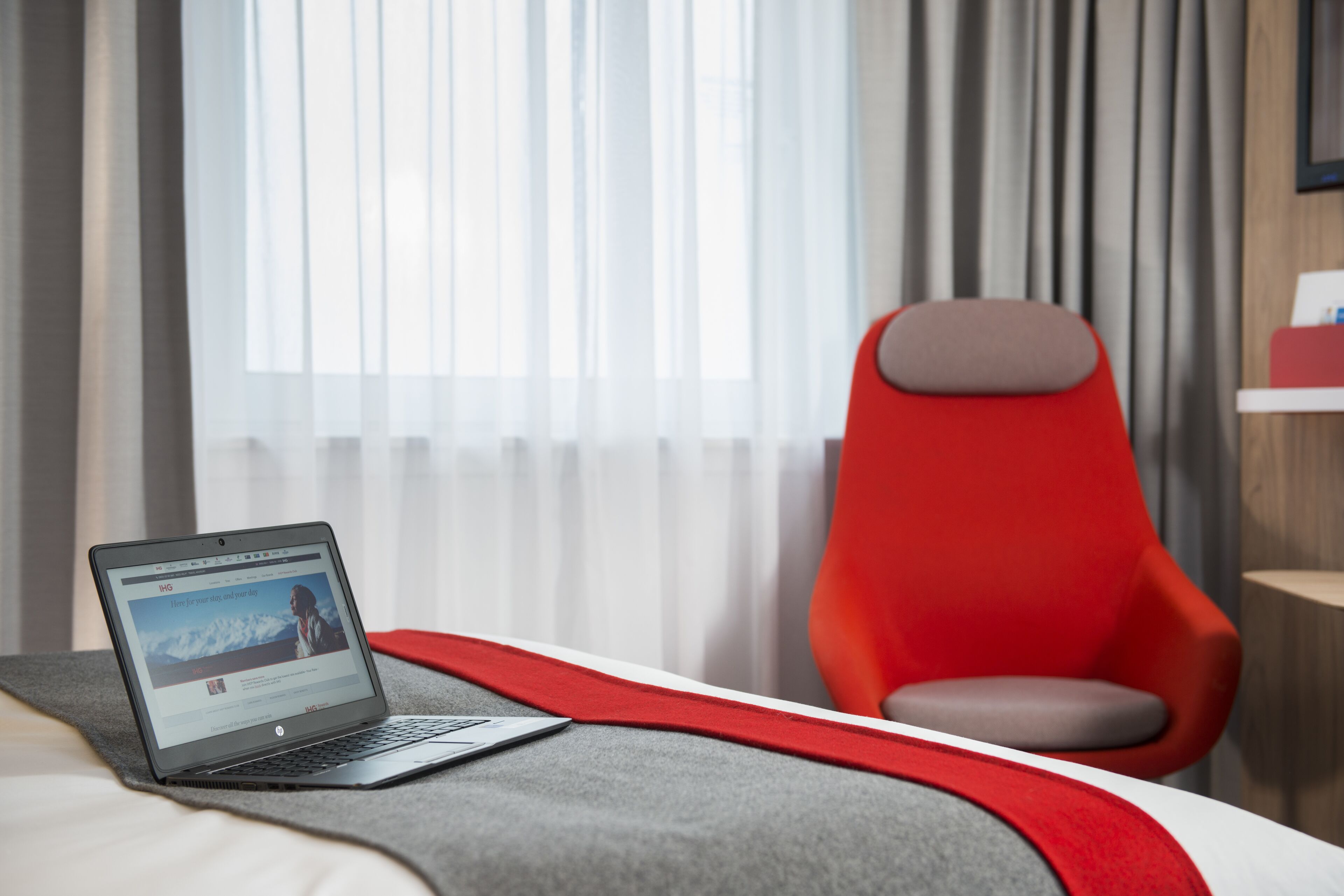Una computadora portátil sobre una cama, con una silla roja y una ventana con cortinas.