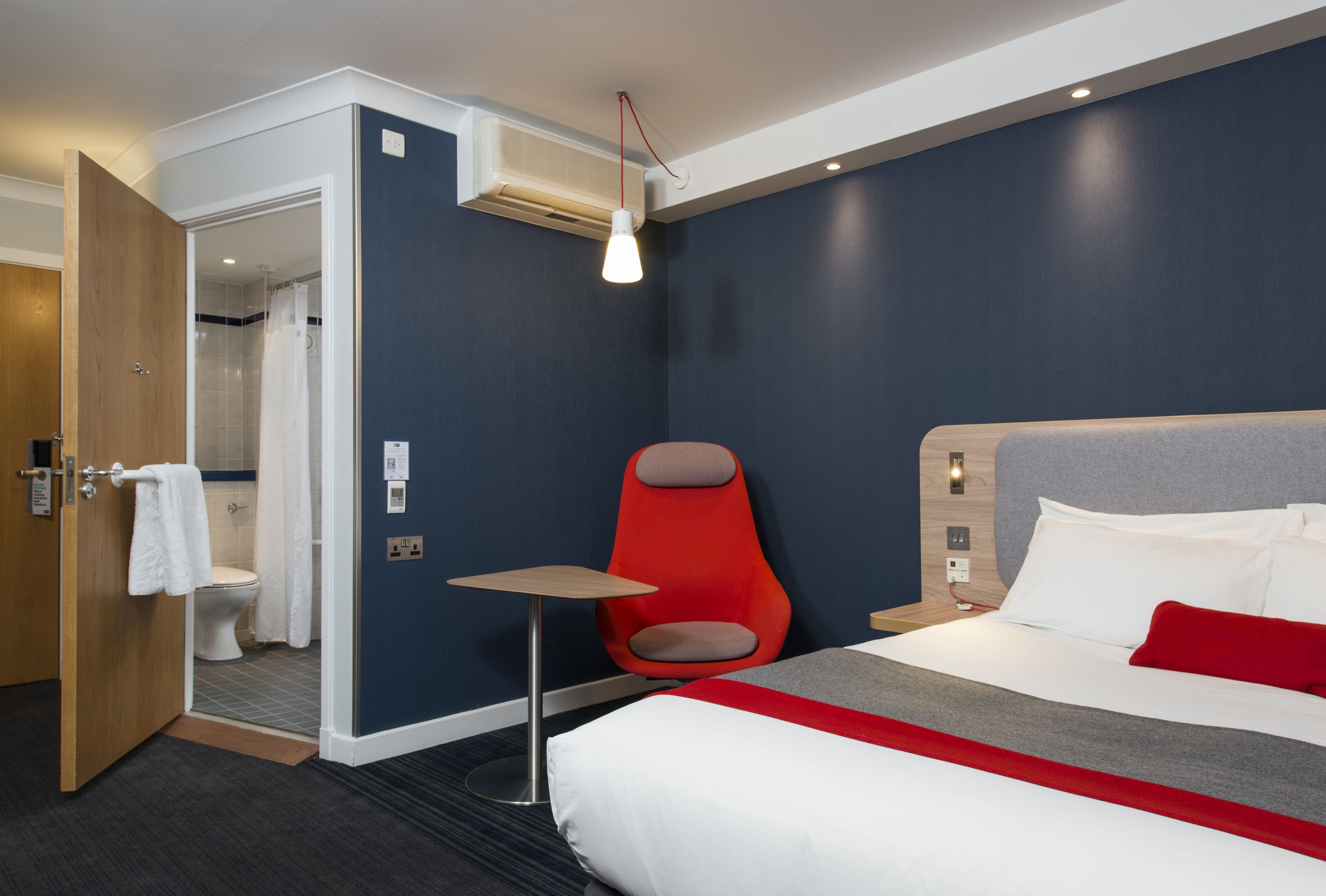 A hotel room with a bed, a red chair, a small table, a shower area, and a door.