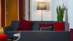 A modern lounge area with a grey sofa, red and grey cushions, a round wooden table, and a red ottoman. - Holiday Inn Express Bath by IHG (Bath)