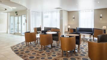 A hotel lobby with a seating area featuring sofas and tables, a patterned carpet, and a large window with curtains.