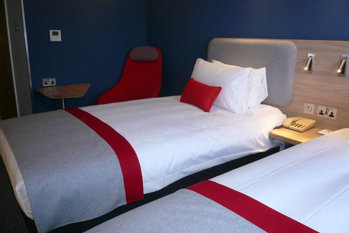 A hotel room with two beds, a red headboard, a bedside table with a telephone, and a wall-mounted light switch.
